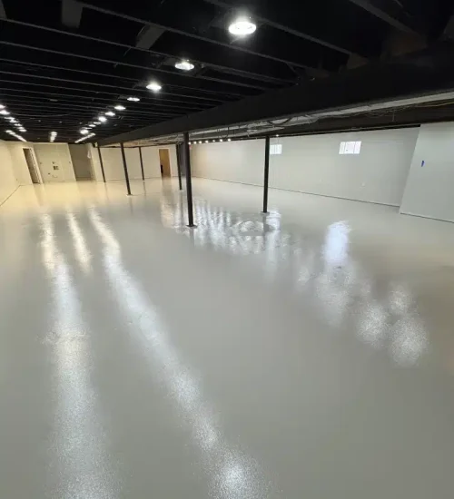Basement epoxy flooring with a glossy light gray finish, white walls and exposed black ceiling beams.